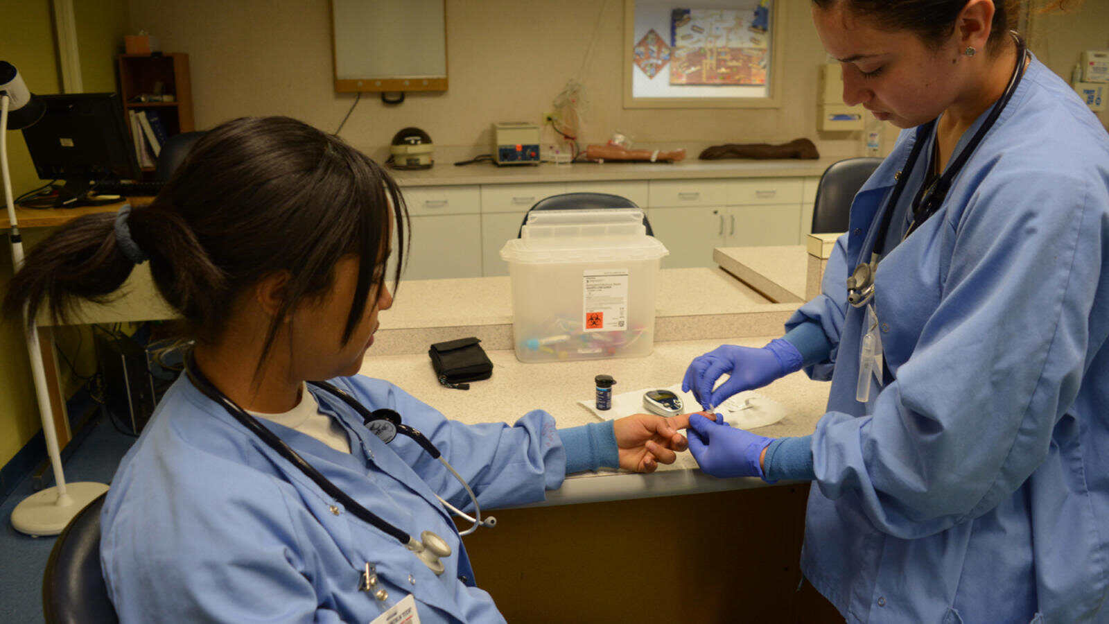 Two medical assistant students practice fingertip blood glucose serum testing.